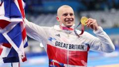 adam peaty holding up a gold olympic medal while wearing his great britain gear