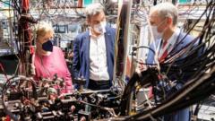 Angela Merkel stands next to two men in front of a complex, somewhat messy array of pipes and wires