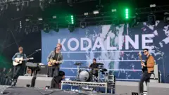 Kodaline play on stage. There is a banner behind them which says 'Kodaline'.