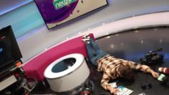 Presenter Ricky Boleto lying face down in the Newsround studio. Various broadcast equipment is scattered on the floor.