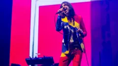 BBC A man wearing a multi coloured jacket, shirt and trousers, sings into a microphone while performing on stage.