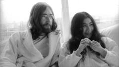 Fotografia em preto e branco mostra John Lennon, um homem branco de cabelos no ombro, barba, e óculos redondos, ao lado de Yoko Ono, uma mulher japonesa de cabelos compridos; ambos usam roupas claras confortáveis e estão sentados em um sofá em frente a uma janela