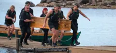 Contestants on the traitors carrying a coffin along a platform