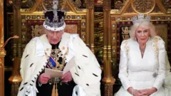 The King and Queen are sitting in large gold chairs. They are both wearing crowns. King Charles is addressing MPs and is reading a speech from a book in his hands. 