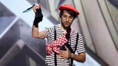 Guillaume Broche, wearing a red beret, striped t-shirt plus red and white checked sash around his shoulder. He's standing behind two microphones, delivering a speech to the audience.