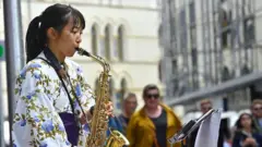 A girl wearing a white kimono with a pattern of blue flowers and green leaves plays a saxophone. In the background, people are walking along a city street. 