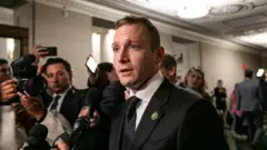 Max Miller, wearing a suit and tie, speaking to reporters holding up microphones in a capitol building