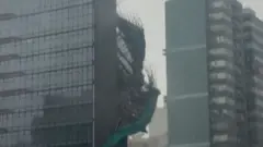 Bamboo and green plastic sheeting are seen falling down the side of a glass skyscraper.