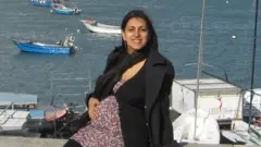 Geeta is heavily pregnant, sat outdoors with the coast behind her and small boats on the water. She is wearing a floral long top, with black trousers, cardigan and jacket and has her hand on her bump.