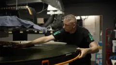 A man with silver hair and black T-shirt leans across an enormous reel of film