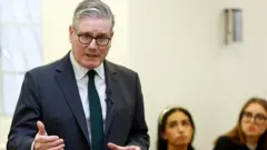 Keir Starmer, in a suit and tie, speaking while two women in the background watch on
