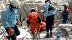 Workers wearing blue and orange PPE can be seen amid the rubble of a building.
