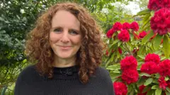 BBC/Katie Razzall Maggie O'Farrell with curly auburn hair in black jumper. Background shows leaves on left and red flowers on the right