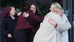 PA Media Two pairs of women embrace and a fifth one looks on. The nearest couple are both wearing cream coats and have blonde hair. Next to him are two men wearing maroon coats and black hair. The woman standing nearby has dyed red hair and is wearing black clothes. They are all smiling happily.
