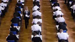 High-angle view of students in uniform sitting at individual desks in rows on a herringbone wooden floor during an exam, while an invigilator walks down the aisle.