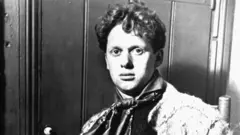 Getty Images/Hulton Archive A black-and-white photograph of a young Dylan Thomas, wearing a cravat and looking directly at the camera. He is sitting down and has short curly hair.