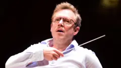 Daniel Harding in an open-necked white shirt with an arm raised holding a conductor's baton conducting the Orchestra Santa Cecilia of Roma in Bologna on 8 May 2026