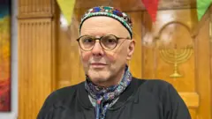 Rabbi René Pfertzel is stood in front of a wooden case and is wearing a neck tie and a yarmulkes as he stands in the synagogue.