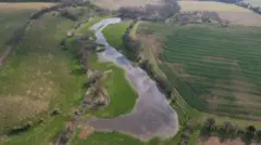 An image captured by a drone shows a river winding through arable farmland.  The river has burst its banks in places and much of the neighbouring field is underwater. 