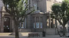 The entrance to the Jersey's Royal Court on a sunny day. In the paved area leading up to the door there are two trees, a bench and a bin. There are three steps leading up to the door which is brown wood and glass. The granite building is ornate with columns and elaborate block work.