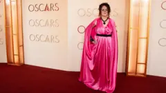 Getty Images Maggie is bright pink long satin dress, black belt, black necklace & little veil hat.
Posing behind the word Oscars.