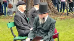 A man with a black top hat and dark suit with a long beard. Her is sat on a chair reading a brown leatherbound book. Behind him are another to men in dark suits and hats on a green painted wooden bench on a green in front of a tree.