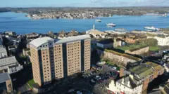 A drone image of the existing building which is set to be replaced. It is a large brown high rise 14-storey building with grey roof. It towers over the buildings set around it. A car park wraps around the building.Plymouth Sound is a short distance away and buildings can be seen on the opposite back. There are three ferries crossing the water. 