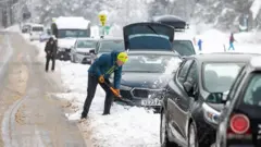PA Media A man removes snow from his car as heavy snowfall causes disruption to roads