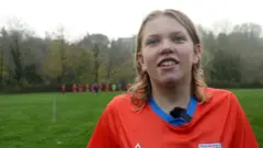 Twelve-year-old BBC Young Reporter winner Alice, wearing an orange football kit with her team in the distance behind her