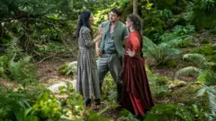 Universal Pictures [L-R] Behind-the-scenes image showing director Chloé Zhao on the set of Hamnet with Paul Mescal and Jessie Buckley
They are standing in the middle of a lush green forest.
Chloe with long black hair in long checked grey dress
Paul in moss green top and trousers
Jessie in dark red dress