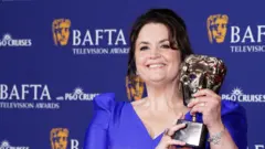 PA Media Ruth Jones holds a BAFTA award in the shape of a theater mask, with a purple background behind it. She is wearing a blue dress with mid-length sleeves and has tied up her black hair.