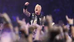 Lead singer and guitarist of Metallica, James Hetfield performs onstage at Lumen Field. He's surrounded by a crowd of people with their arms up. He's wearing a black t shirt, arm cuff and a denim vest. He's holding a guitar. He's got his arm up in the air and appears to be shouting.
