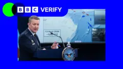 Gen Dan McCaine wearing a blue US Air Force uniform stands at a lectern in front of a screen showing a map of the Strait of Hormuz and the Gulf of Oman