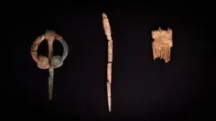 Kevin Church/BBC News Three artefacts are displayed against a black background. On the left, is a green almost circular metal brooch with a long pin. It has tiny dots etched into the pin, while one of the terminals of the brooch has four large dots. It is partially covered in soil.
In the centre of the picture is a decorated pin made of bone. It is broken into four pieces. The top of the pin is intricately carved with a pattern. On the right is a fragment of a comb with broken teeth that has been carved from antler.