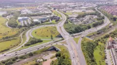 Ariel photograph of the large A135/A66 junction on the South Stockton Bypass showing moving traffic and curved duel carriageways weaving through suburban and industrial areas and grassy embankments. 