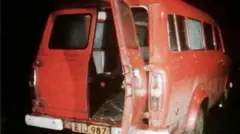 Rear shot of a red mini bus with back door lying open, windows on the van are smashed