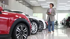 Getty Images A woman wearing a gray jacket and blue jeans walks through a bright white car showroom and looks at a row of vehicles.