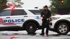 A police officer with a gun stands in front of two white marked police cars
