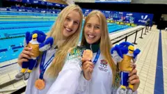 Nel family Olivia (left) and Georgia Nel stand in front of a swimming pool. They both have long blonde hair and are wearing white tracksuits, carrying toy birds and wearing gold medals.