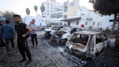 People inspect Al-Ahli hospital with burned out vehicles in the foreground