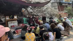 A group of people sits on the ground crossed by legs, turning to several monks who sat in the chairs who turn to them - all in front of the partially destroyed building