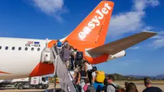 Getty Images Some people aboard an EasyJet flight are carrying hand luggage and rucksacks on the tarmac of the airport in Sardinia. It is a sunny day and the sky is blue.