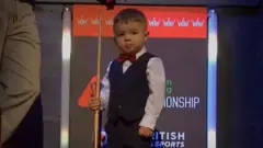Luke stands on a stage wearing a black waistcoat over a white shirt. He wears a red bowtie and holds a small snooker cue