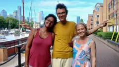 Tunde, Buddy and Kellie stand in a row on a path by a river. Boats can be seen along the river beside them and there are skyscrapers in the distance.
Tunde has short, dark curly hair and is wearing a red strappy top with grey trousers. She has her arm around Buddy's waist. Buddy has dark curly hair and glasses. He is wearing a yellow t-shirt with beige shorts and has his arms outstretched with his index fingers pointing. Kellie has blonde hair and is wearing a colourful patterned summer dress with an abstract design.