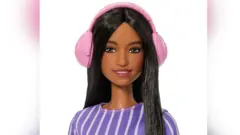 Mattel/PA image of the new autistic Barbie doll. She has long brown hair and is smiling. She is wearing a set of pink ear defenders that look like large headphones and each sits over her ears.