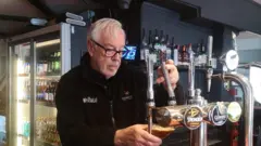 Geoff Robbins An old man in black wool with the pub branding in white is pulling a pint behind the bar in a pub. In the background is a fridge with interior lights containing beer bottles.