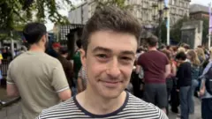 A man wearing a navy and white stripped t shirt is seen smiling at the camera as he stands in front of the city hall in Belfast ahead of the start of the Fleadh Cheoil na hÉireann with several dozen other people standing in the background.