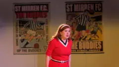 A woman with red hair and a red/pink dress, standing on stage in front of two posters showing support for St Mirren football club.