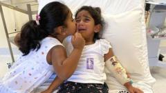 Tharnicaa crying on a hospital bed as her sister Kopika kisses her cheek to comfort her