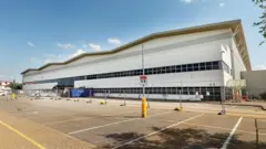 A large white warehouse building with black windows. There are fences in front of the building and an empty car park.